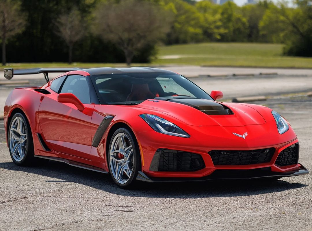 A red sports car with black accents and a rear wing, one of the Top 10 American Rides, is parked on an empty asphalt lot with trees and grass in the background.