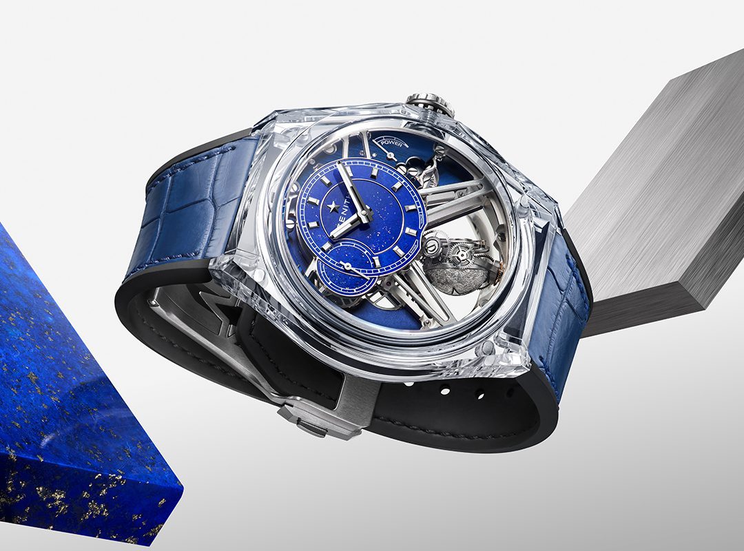 A luxury Zenith DEFY Zero G wristwatch with a blue dial, visible mechanical parts, and a blue leather strap from the Sapphire Collection, displayed on geometric blocks against a white background.
