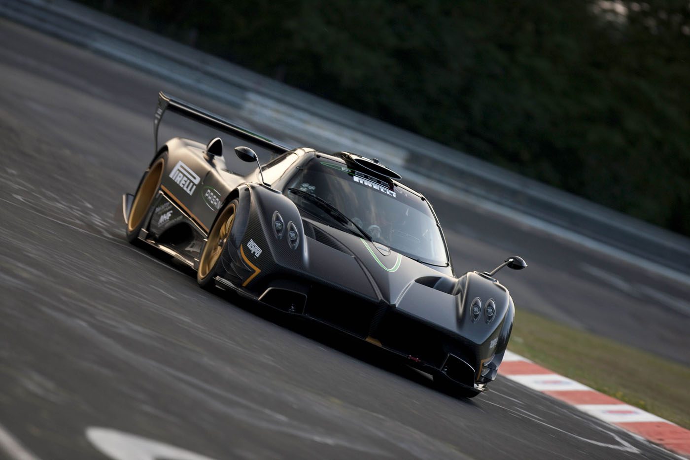 A black Pagani sports car with gold accents speeds through a racetrack corner at 2025 Monterey Car Week, slightly tilted as blurred trees and barriers rush by in the background.