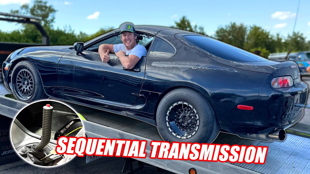 A man leans out of the window of a dark Toyota Supra sports car on a trailer; inset shows a Sequential Gearbox shifter. Text reads "SEQUENTIAL TRANSMISSION.