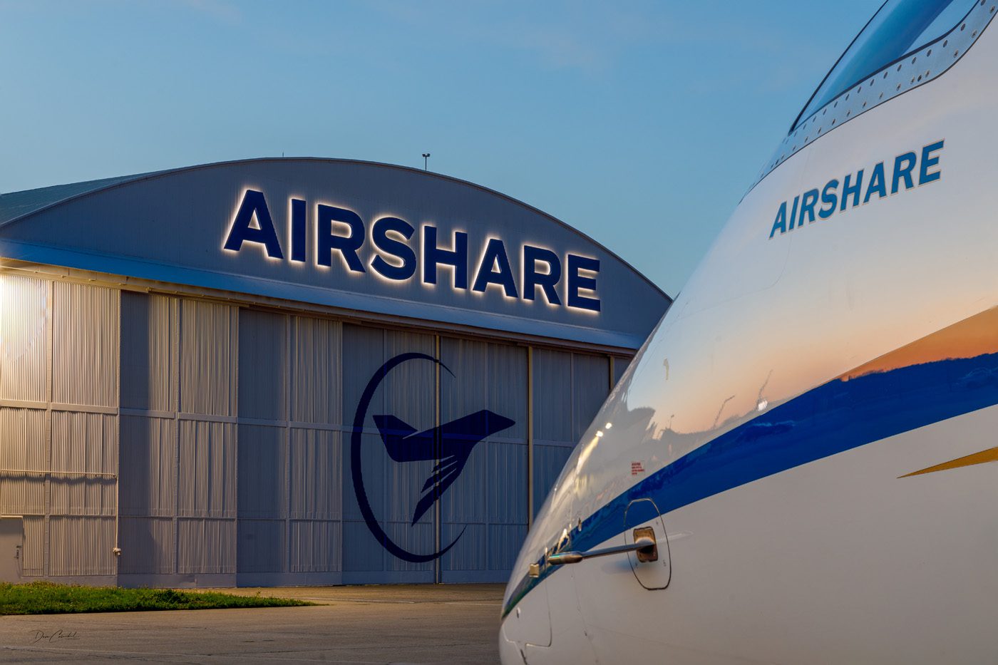 Aircraft parked outside an Airshare hangar at dusk, with the hangar logo illuminated. The plane's nose peeks into view, embodying a perfect fit against the evening sky, ready to fly.