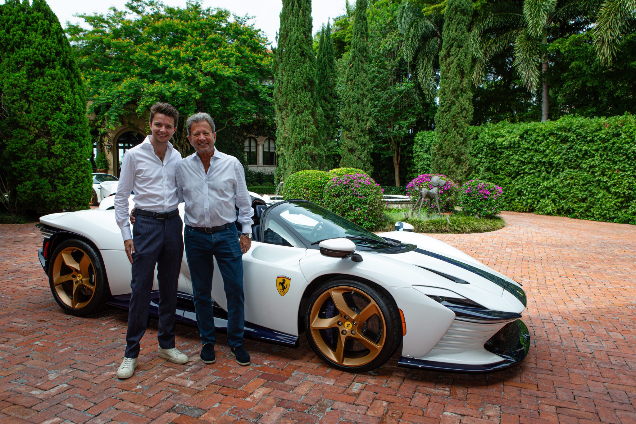 Two men stand smiling next to a white Ferrari sports car with gold wheels, parked on a brick driveway surrounded by greenery—an elegant scene reminiscent of a fine art collection.