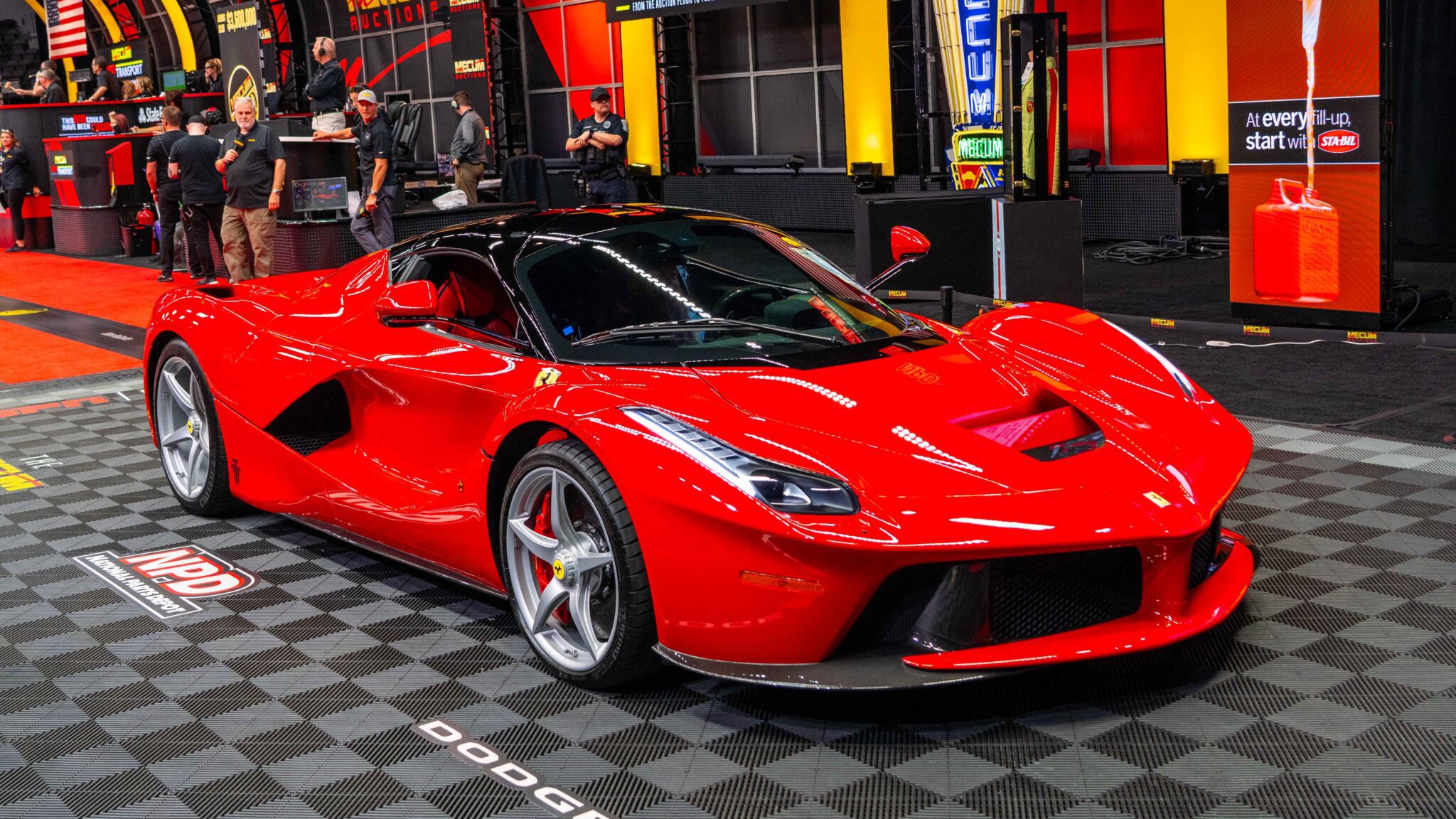 A red sports car is displayed indoors on a checkered floor, with several people and automotive displays in the background.