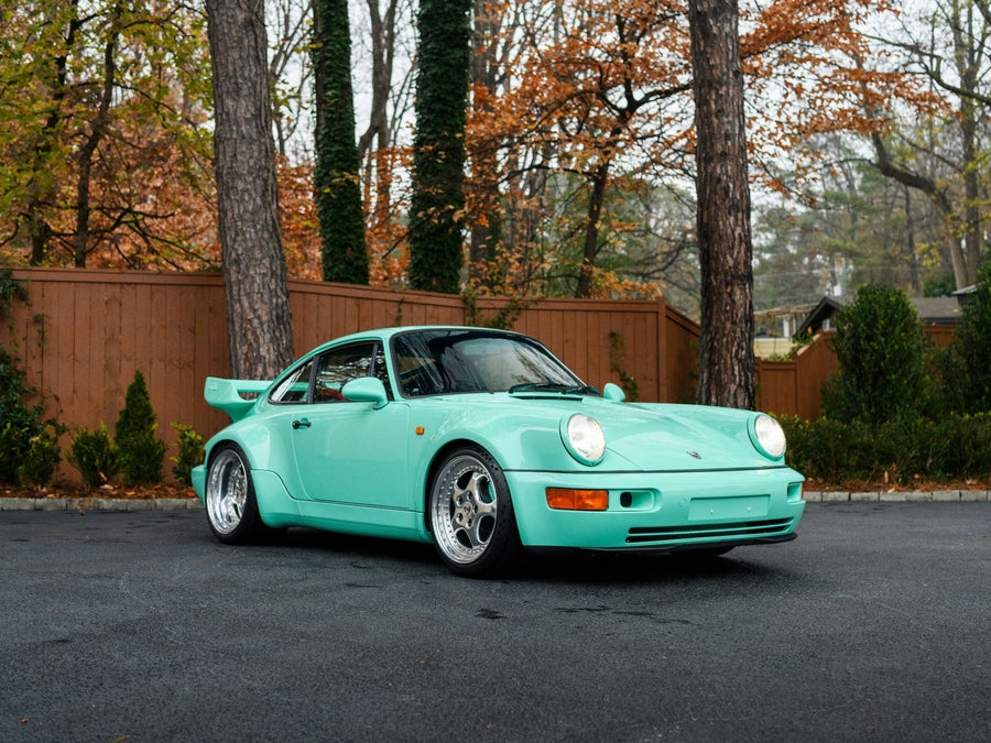 A mint green Porsche sports car from Merit Partners' exclusive selection is parked on a paved driveway, surrounded by tall trees and a wooden fence with autumn leaves in the background.