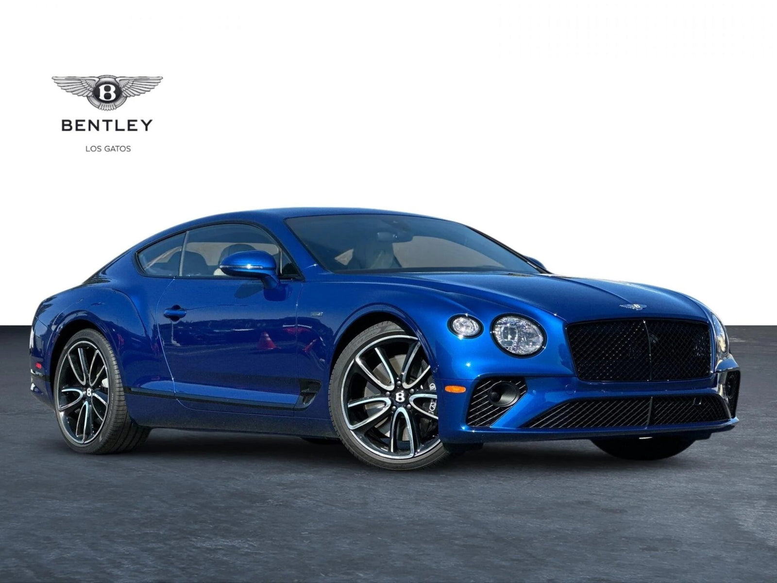 A blue Bentley Continental GT coupe is parked on a paved surface with the Bentley Los Gatos logo in the background.