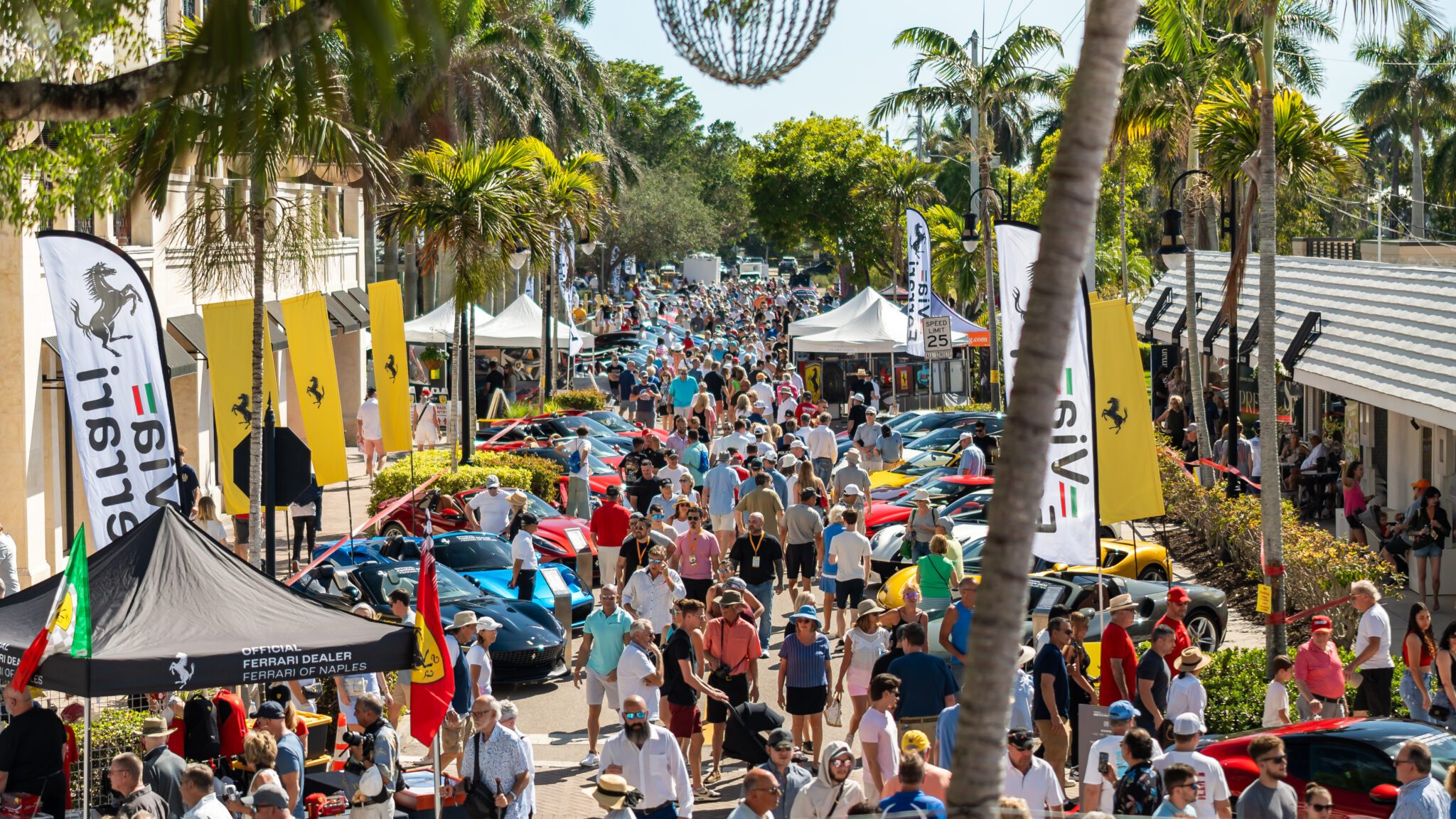 A large crowd walks among rows of parked sports cars at the 2026 Concours Naples Cars on 5th, an outdoor car show lined with palm trees, vendor tents, and banners on a sunny day.