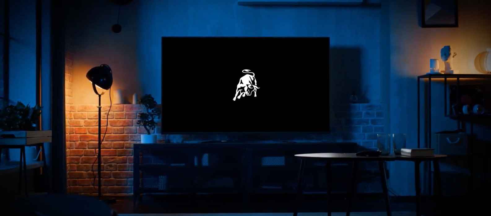 A dimly lit living room features a large Smart TV App displaying a white logo of a bull reminiscent of Lamborghini on a black screen. A floor lamp and assorted furniture are visible, creating an inviting atmosphere for viewers worldwide.