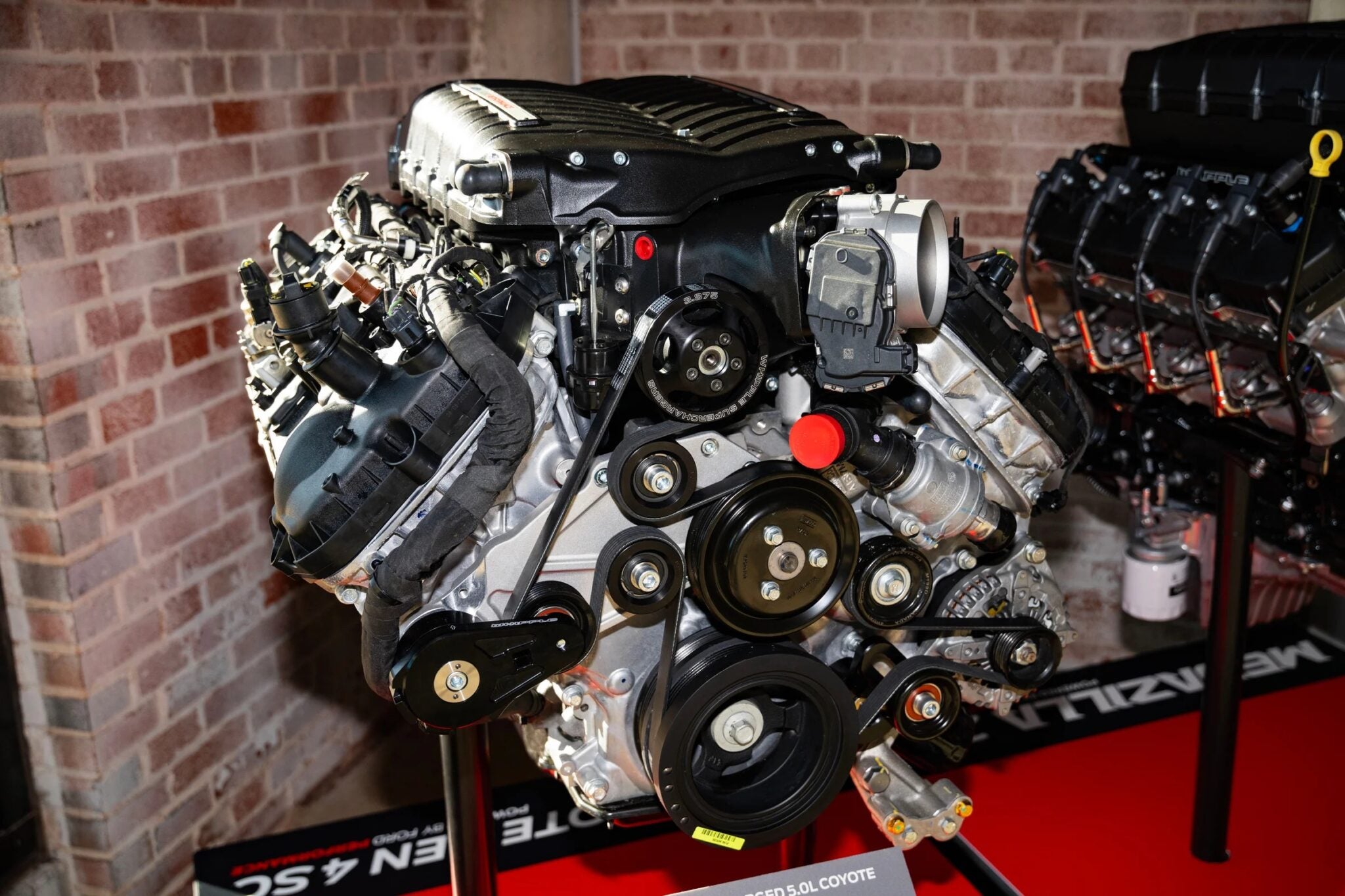 A close-up of a Ford Megazilla engine displayed indoors on a stand, showcasing its 1000+ HP prowess and intricate belts and components against a brick wall backdrop.