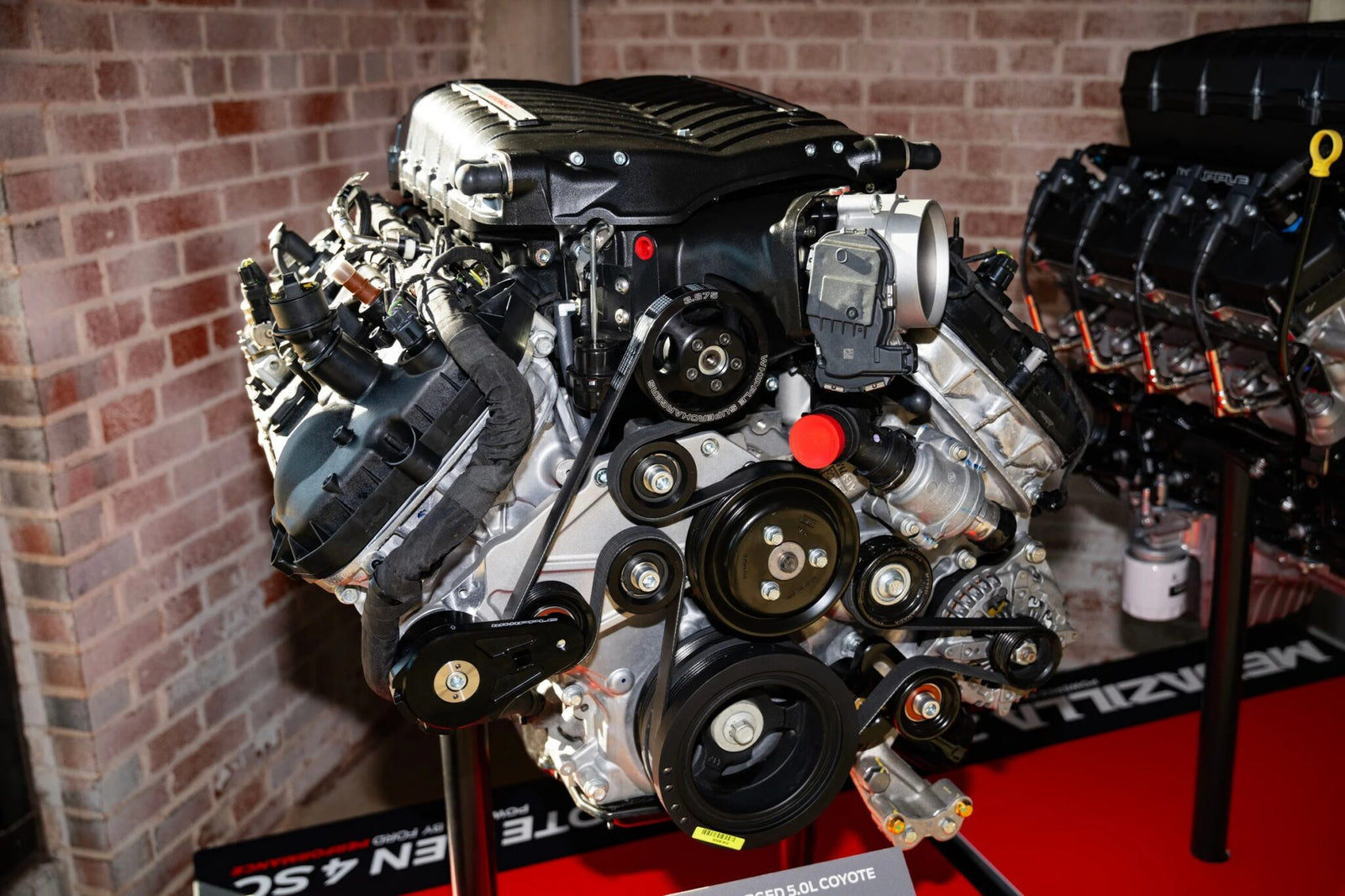 A close-up of a Ford Megazilla engine displayed indoors on a stand, showcasing its 1000+ HP prowess and intricate belts and components against a brick wall backdrop.