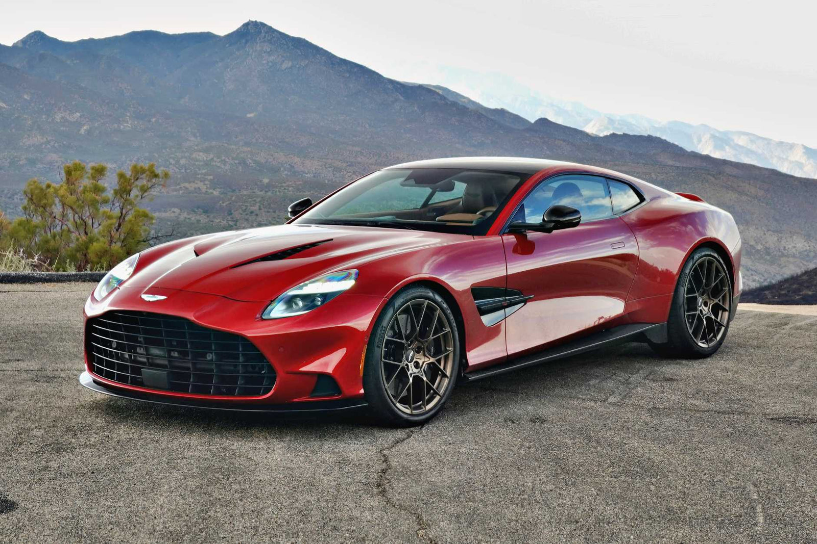 A red 2025 Aston Martin Vanquish Coupe is parked on a paved surface, with mountains and a cloudy sky in the background. Review: This sleek sports car blends elegance and power against a stunning natural backdrop.
