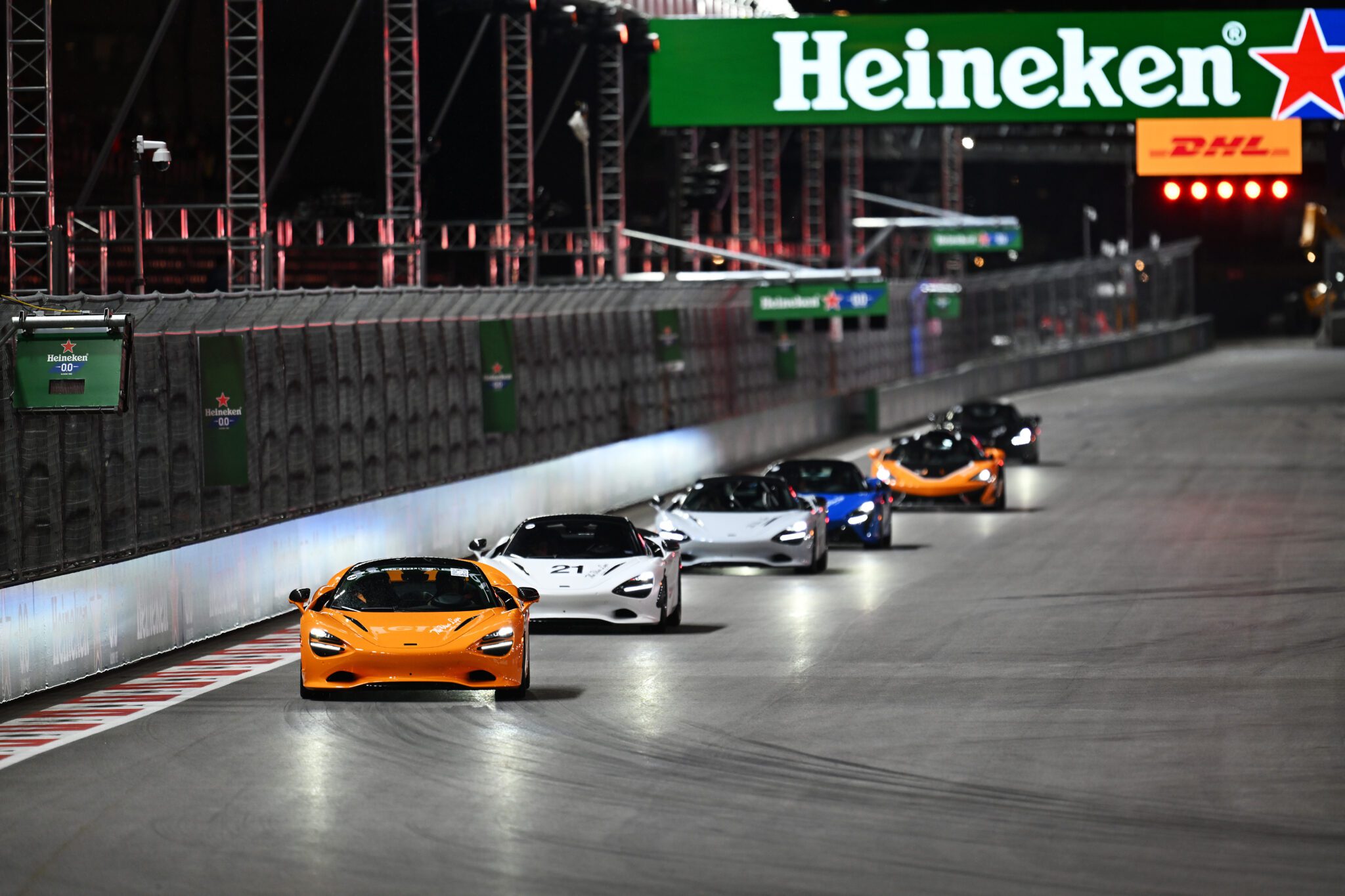 A line of sports cars drives on a nighttime racetrack under a large illuminated Heineken sign, capturing the excitement of Las Vegas F1.