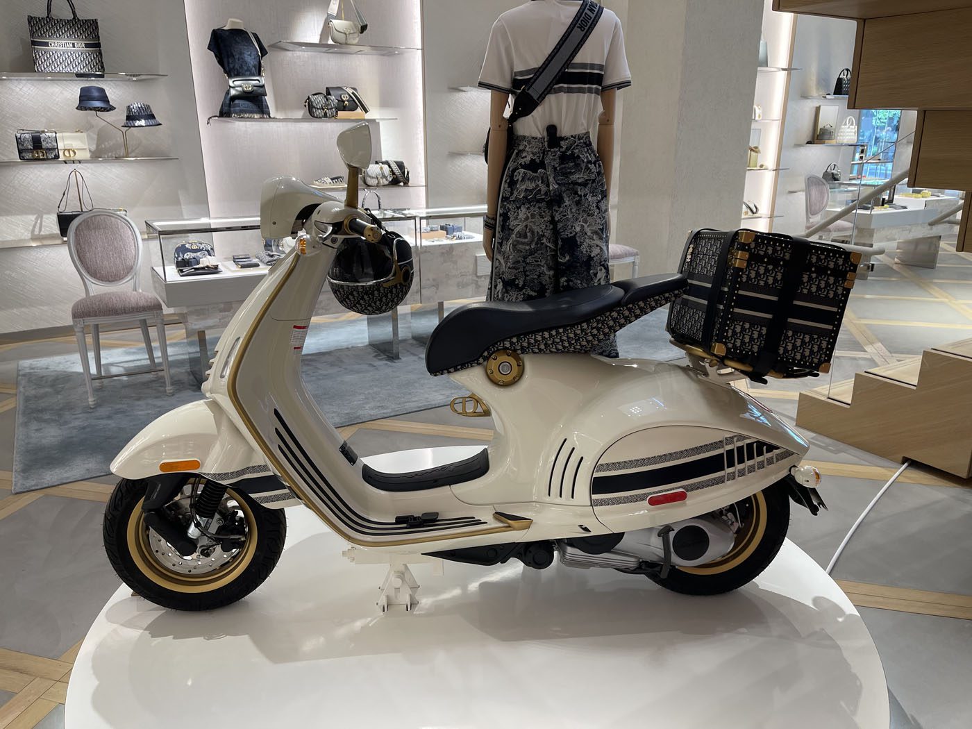 A white and black scooter with a patterned seat and storage box on display in a store. Shopping bags and other items are visible in the background.