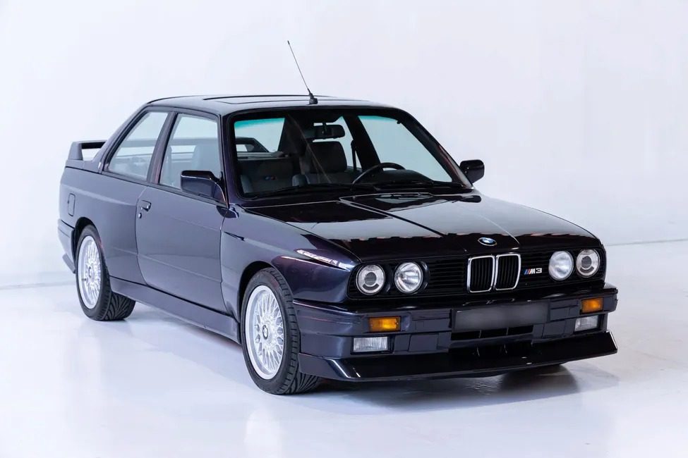 A black classic BMW M3 car, reminiscent of a German heavy metal band's power, is parked in a pristine white studio, viewed from the front-left angle.