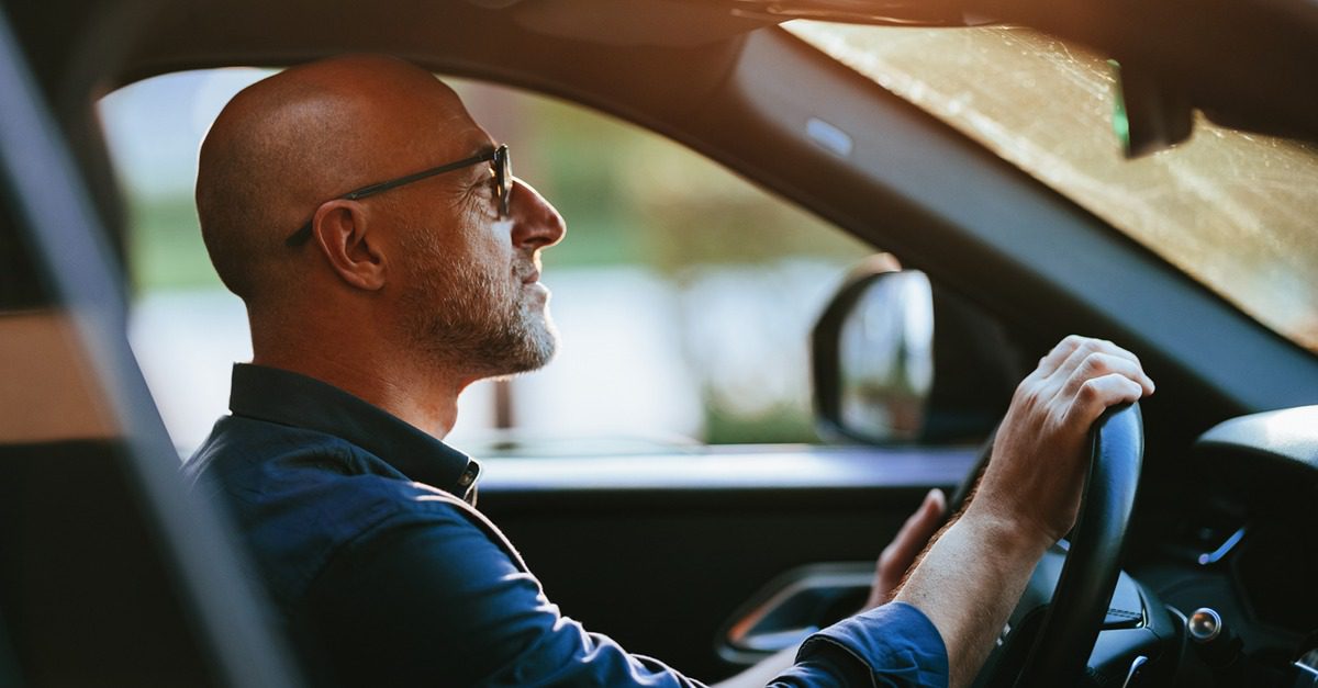 A man with glasses and a shaved head is sitting in the driver’s seat of a car, holding the steering wheel and looking forward, as if preparing to navigate through busy public markets or down Crowd Street.