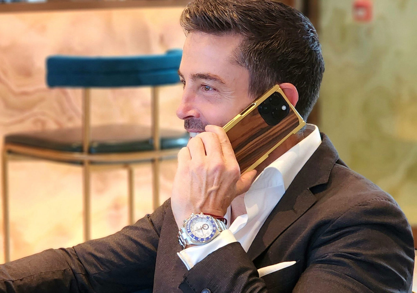 A man in a suit holds a large smartphone to his ear, its elegance elevated by an EXOvault Case. He sits indoors next to a chair, the craftsmanship of his attire matched by his watch.