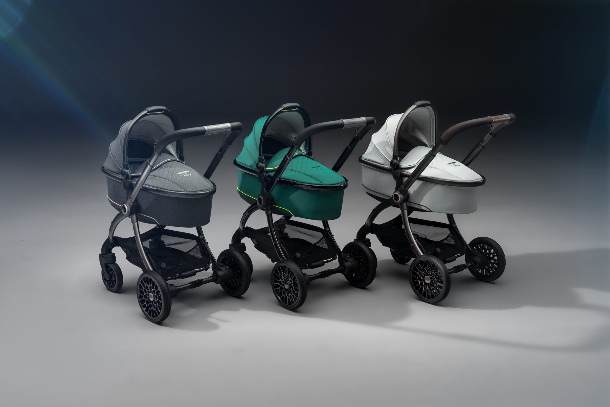Three modern Aston Martin strollers in gray, green, and white are lined up side by side on a plain, softly lit background—perfect for next generation car lovers.