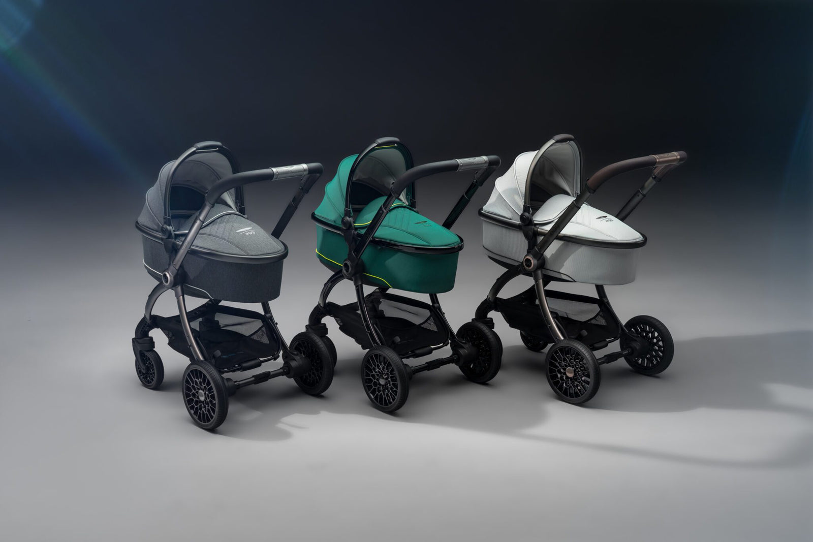 Three modern Aston Martin strollers in gray, green, and white are lined up side by side on a plain, softly lit background—perfect for next generation car lovers.