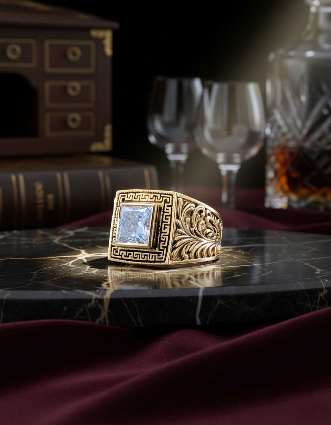 A gold ring with an ornate design and a large square gemstone is displayed on a marble surface, embodying the rare legacies of AviNika Jewelry, with wine glasses, a book, and a decanter in the background.