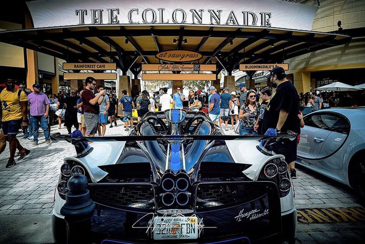 A crowd gathers around luxury sports cars, including a Pagani Huayra, outside The Colonnade shopping area on a sunny day.