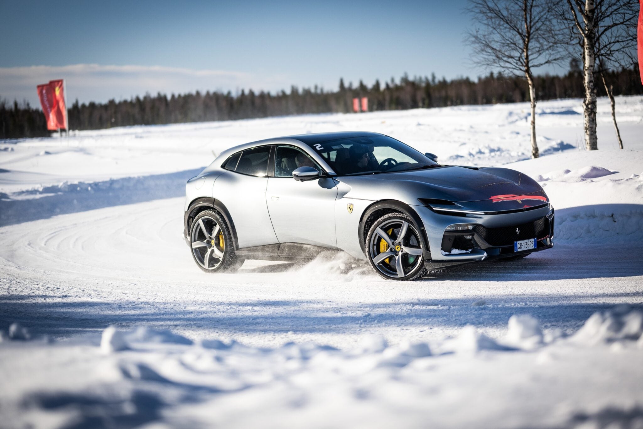 A sleek silver Ferrari 296 GTB navigates a snowy track, kicking up snow. The background features trees and two red flags against a clear sky.