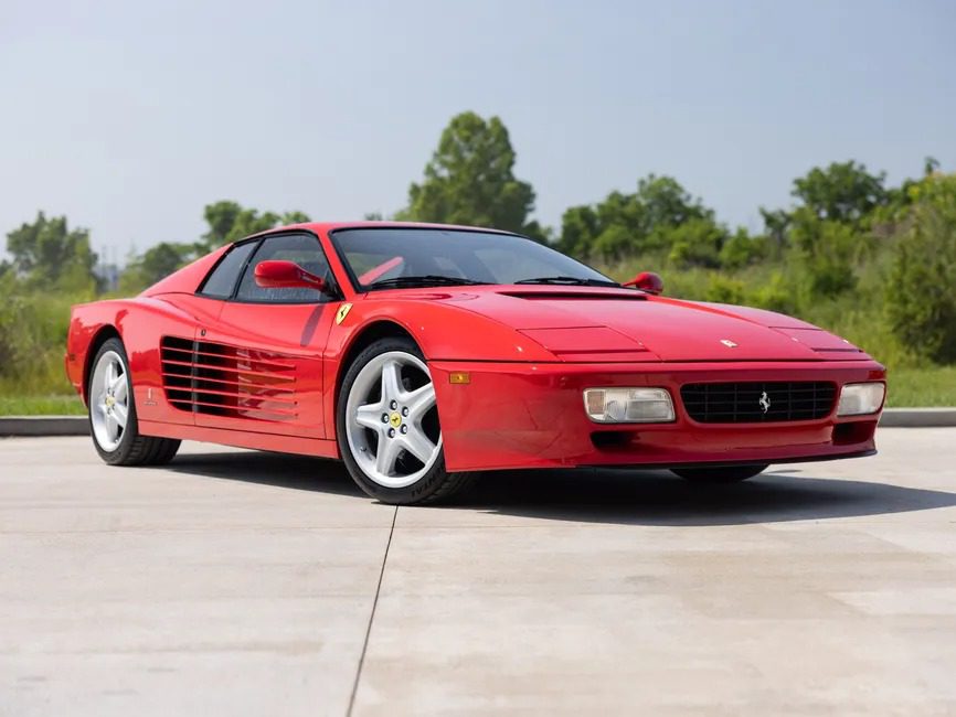A rare Ferrari 512 TR Prototype sports car is parked on a driveway with greenery and trees in the background under a clear sky.
