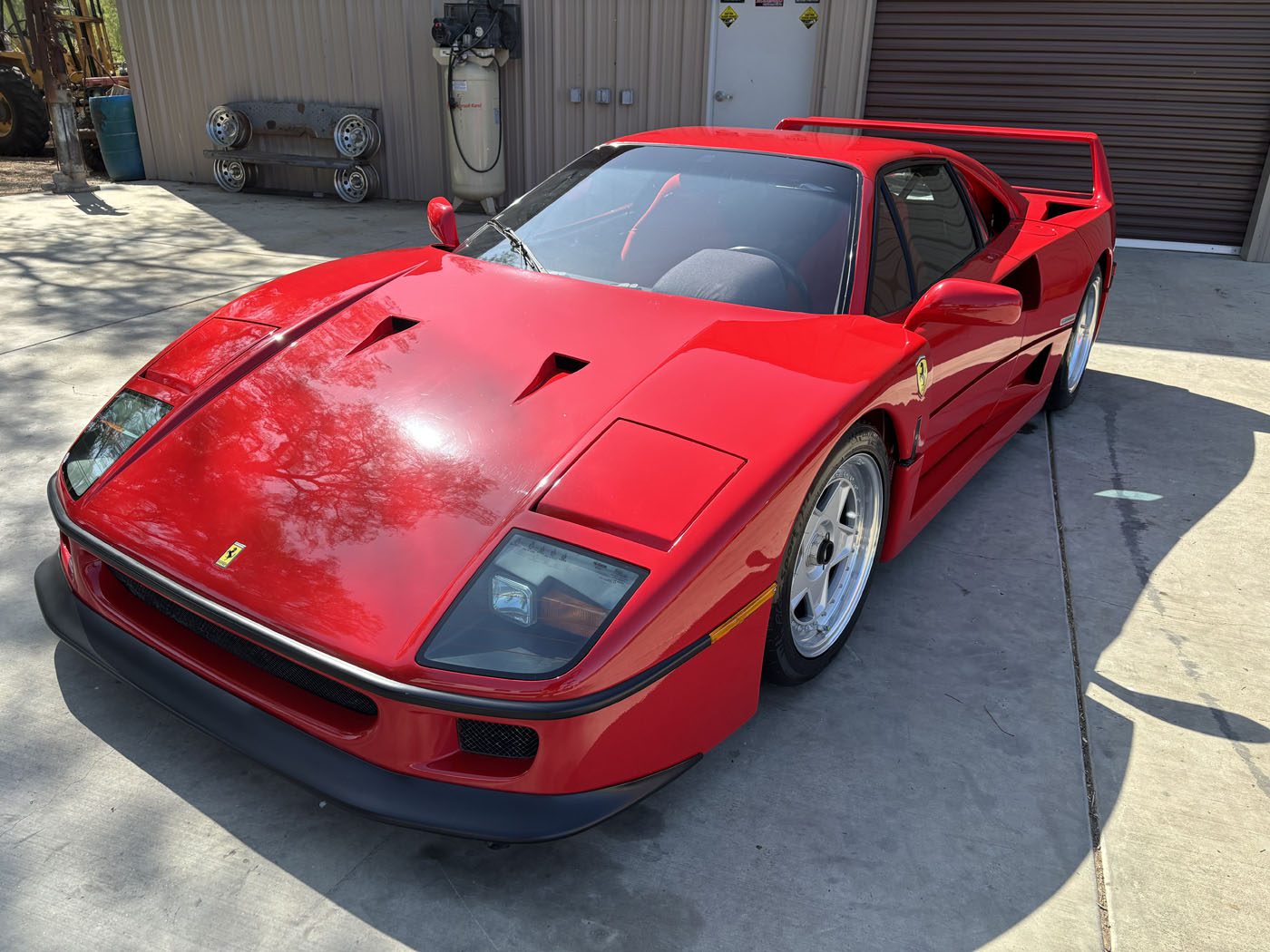 A red Ferrari F40 sports car is parked on a concrete driveway near a building with a closed garage door and equipment mounted on the wall, ready for its next owner—don’t miss your chance to buy it now at auction.