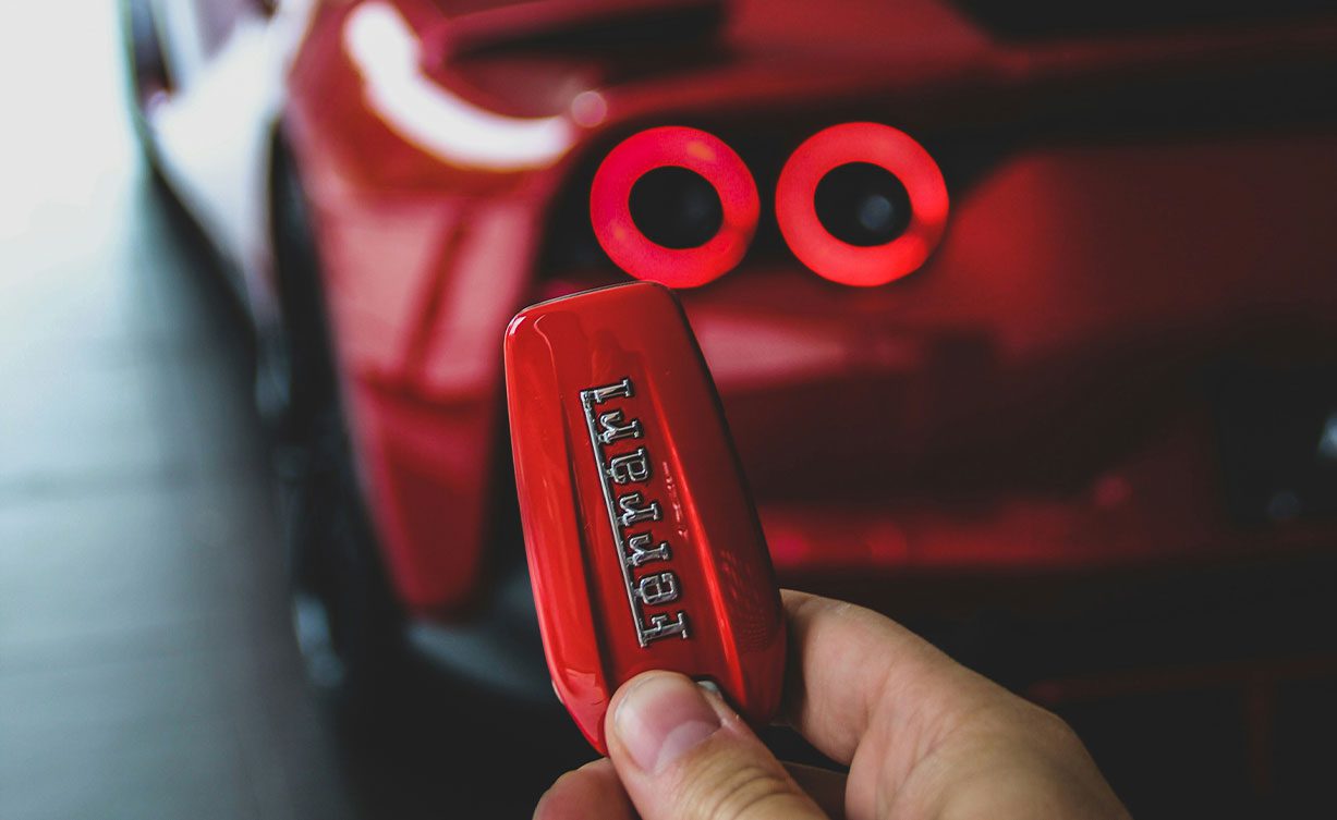 A close-up of a hand holding a red Ferrari key fob, with the blurred rear section of this exotic car showcasing its illuminated tail lights in the background.