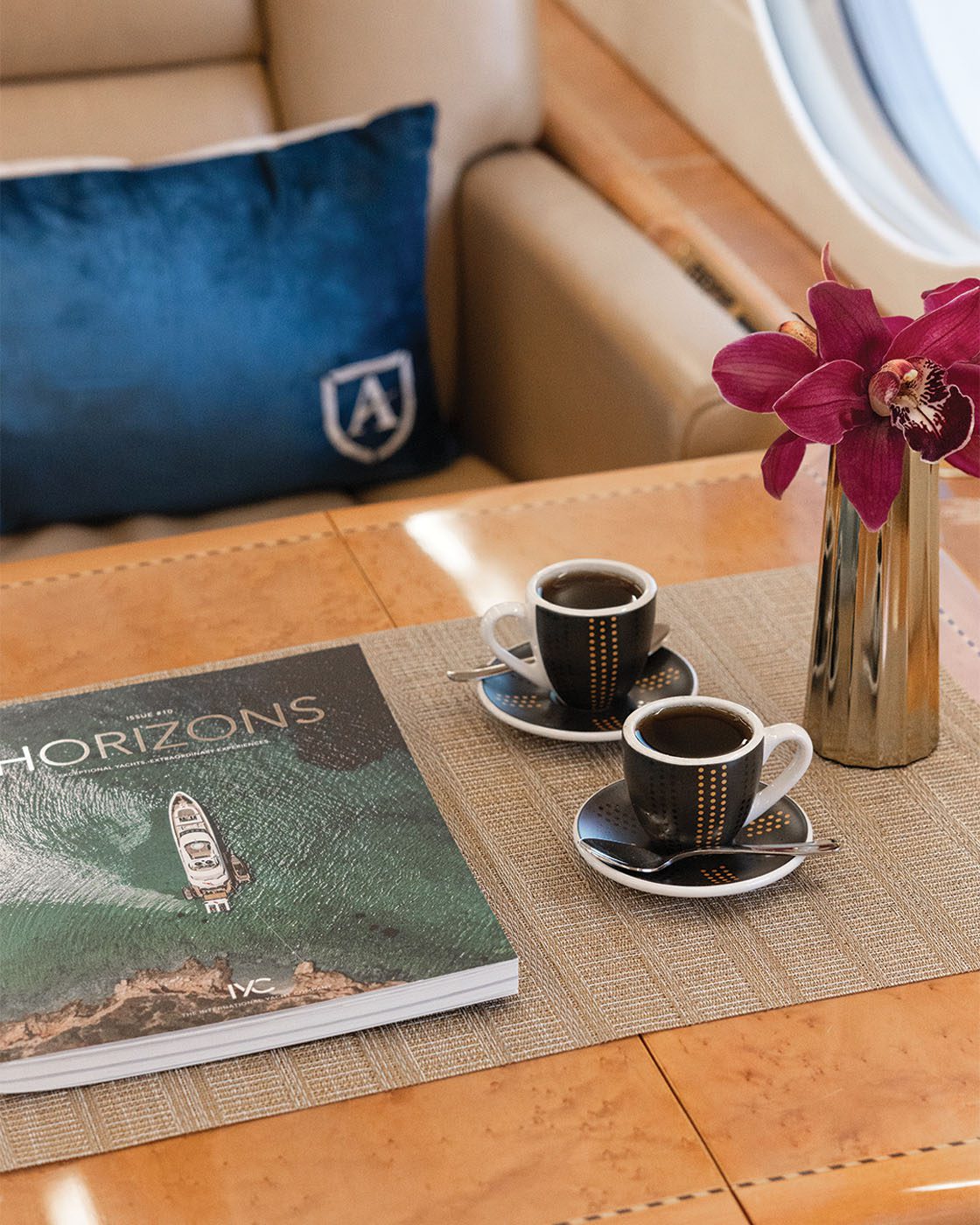 On a table next to a blue pillow, coffee cups sit alongside a book titled "Horizons" and a vase with purple orchids, evoking the elegance of private aviation.