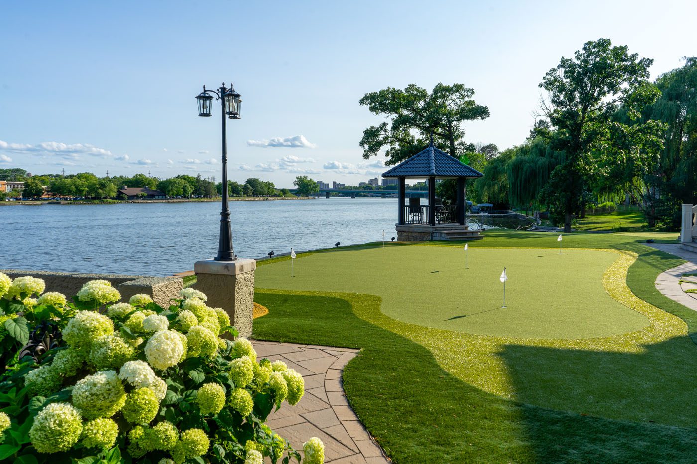 A landscaped backyard features a putting green, a gazebo near the water, flowering hydrangea bushes, and a lamppost under a clear sky.