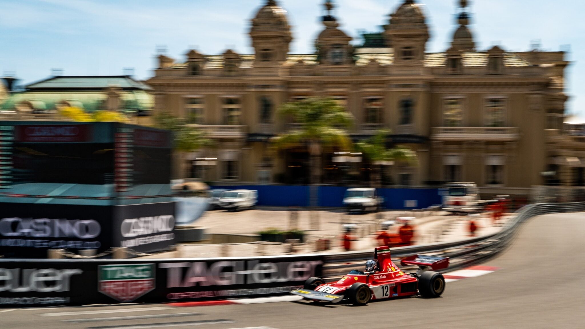 A red Formula 1 car with the number 12 races around a corner on a city circuit during Cavallino Classic Monaco, with an ornate building and palm trees in the background.