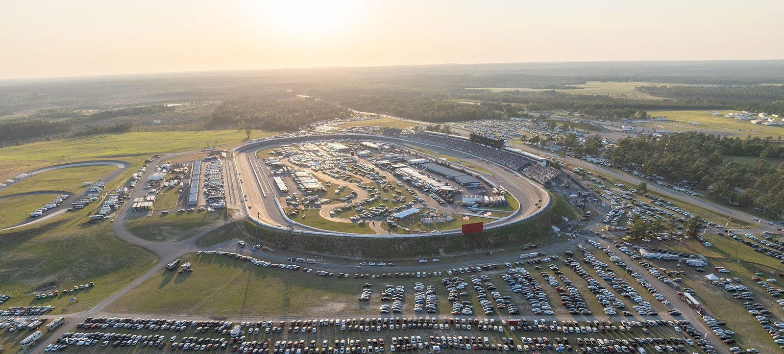 Aerial view of Rockingham Speedway, the ultimate playground for racing fans, surrounded by numerous parked cars and spectators with sunlight illuminating the scene.