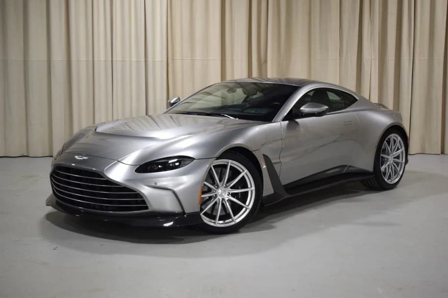 A silver 2023 Aston Martin Vantage coupe is parked indoors on a light-colored floor with beige curtains in the background.