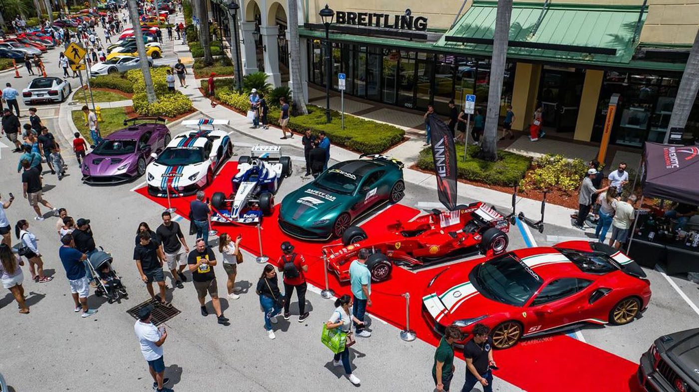 Several sports cars, including a striking Praga Bohema hypercar, are displayed on a red carpet outside a Breitling store as crowds explore this highlight of the South Florida automotive weekend.