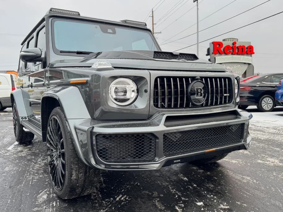 A gray BRABUS-modified Mercedes-AMG G 63 SUV is parked on a snowy lot near a dealership with a red Reina sign in the background, ready for sale.