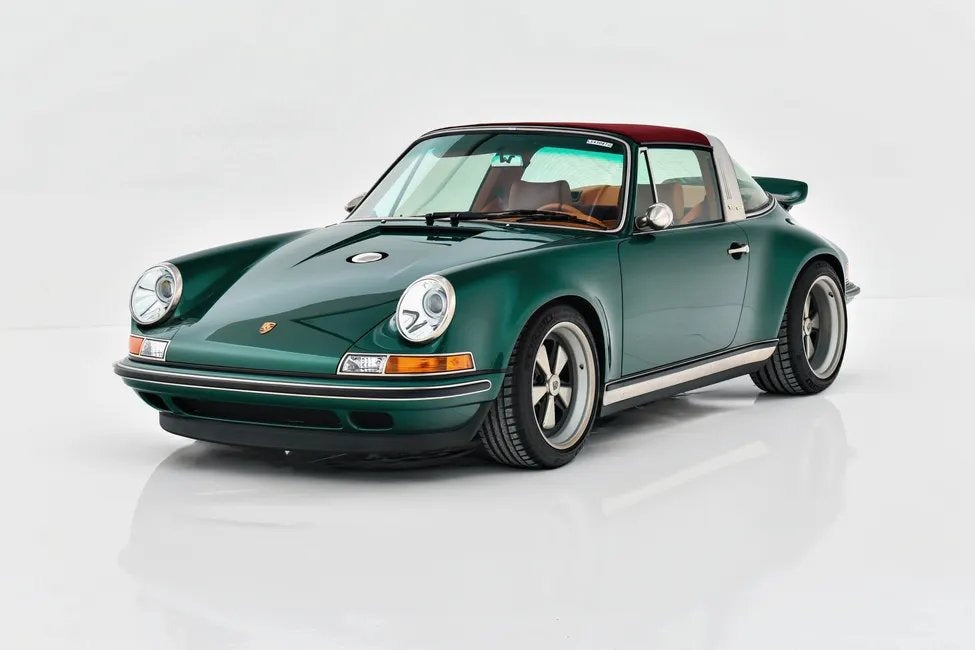 A classic dark green Porsche sports car with a red roof and tan interior is parked on a white reflective surface, viewed from the front left angle.