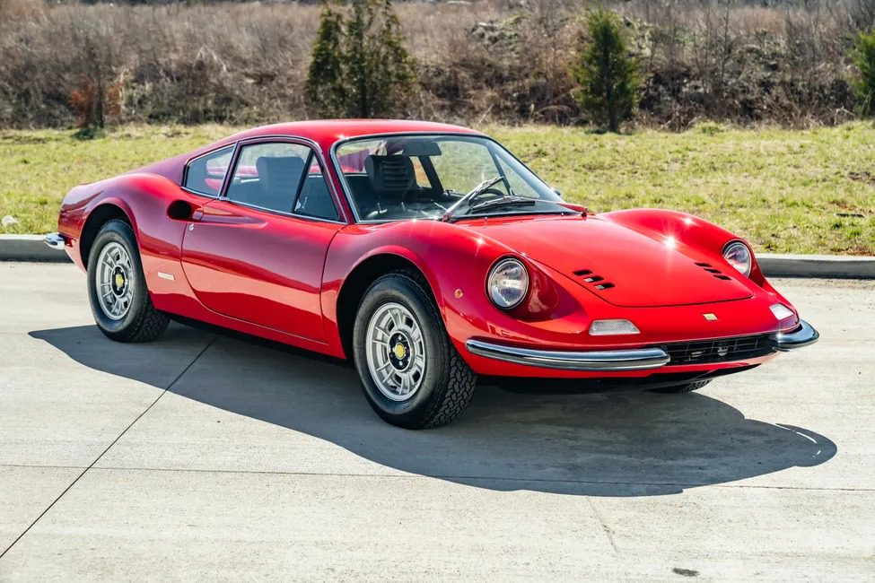 A classic Ferrari Dino 246 GT is elegantly parked on a concrete surface, framed by lush grass and towering trees in the background, hinting at its storied past with Sotheby Motorsport and its allure at auction events.