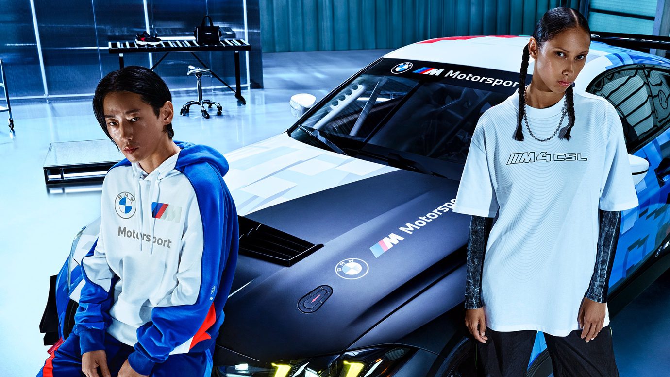 Two people in BMW Motorsport attire stand in front of a black BMW car, showcasing the sleek style of the PUMA x BMW M4-Inspired Collection.