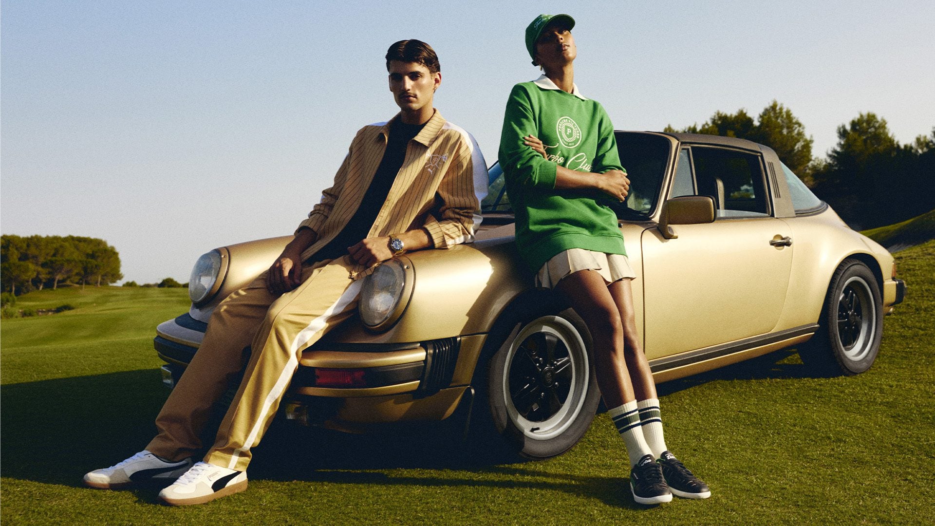 Two individuals lean against a vintage gold car parked on a grassy field. One wears a tan outfit, the other a green sweater and shorts. Trees and clear sky in the background.