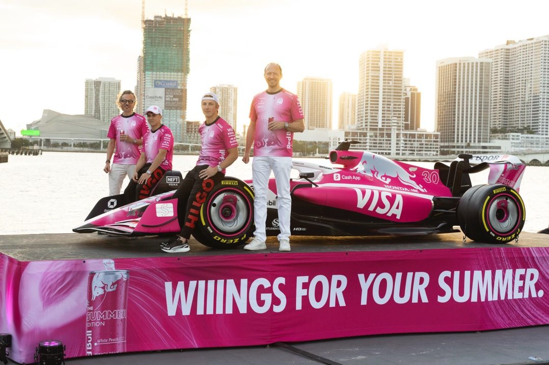 Four people in pink shirts stand beside a pink Formula 1 car with “VISA” branding, displayed on a platform with “WIIINGS FOR YOUR SUMMER” text, cityscape and water in background.