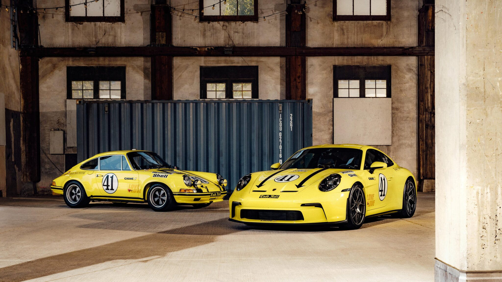 Two yellow Porsche 911 S/T sports cars, each bearing racing number 41 decals, are parked side by side in an industrial warehouse with exposed beams and a large shipping container—a true legend in the world of Porsche 911.