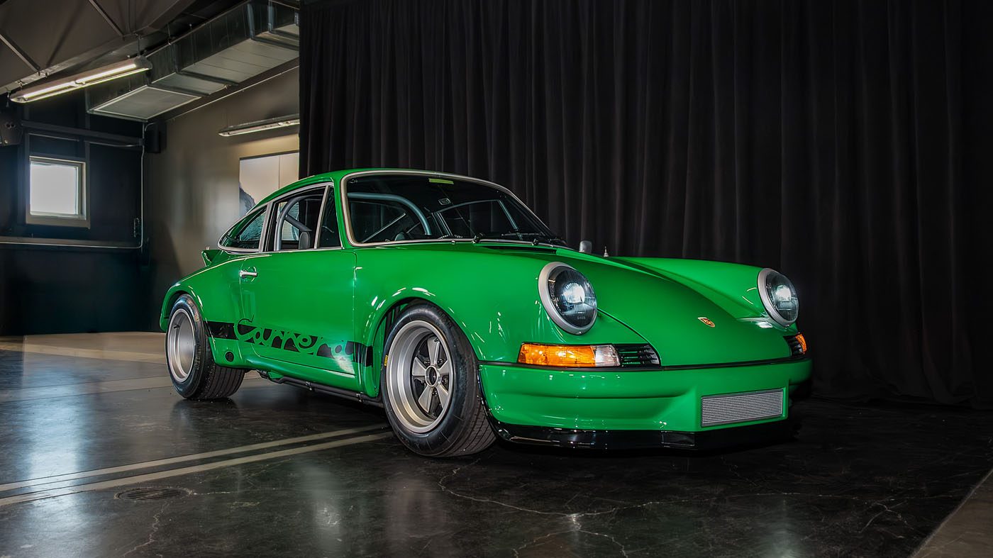 A green 2025 Porsche, a National Winner in Porsche Classic Restoration, is parked indoors on a polished concrete floor in front of a black curtain.