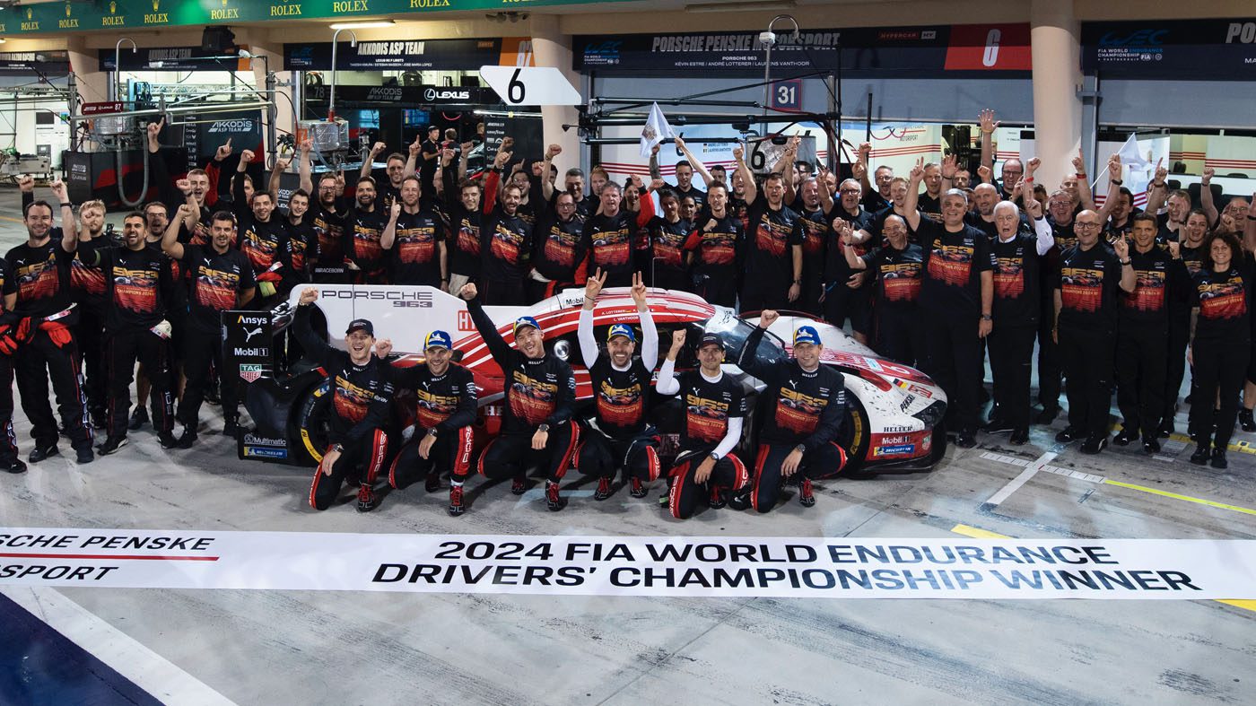 A racing team, comprised of accomplished works drivers, celebrates their FIA WEC 2024 World Endurance Championship win, posing triumphantly around a Porsche with arms raised in a cheering gesture.