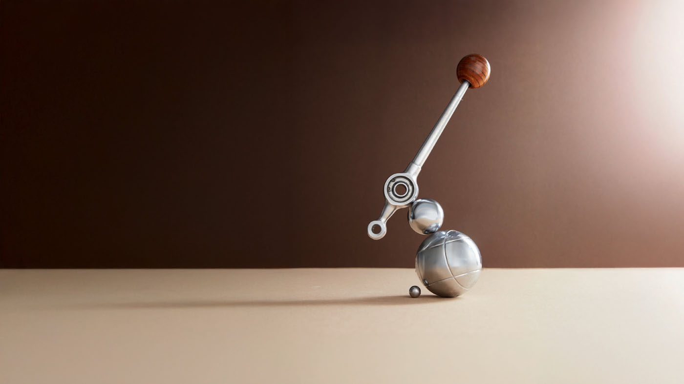 A metallic abstract sculpture with spheres and a lever-like arm topped with a wooden ball, placed on a beige surface against a brown gradient background.
