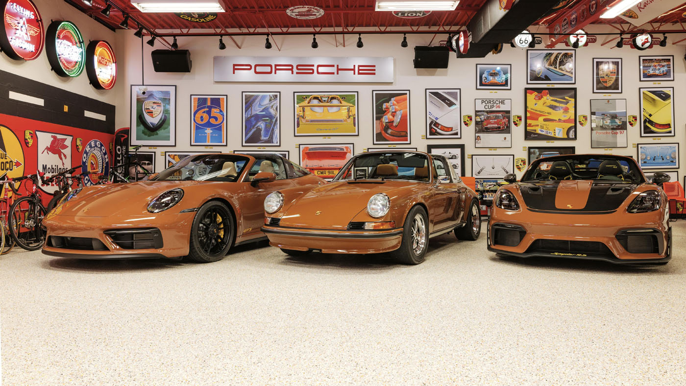 Three Sepia Brown Porsches, celebrating 50 Years of automotive excellence, are parked in a garage filled with collector memorabilia, car-themed posters, and vibrant neon signs.