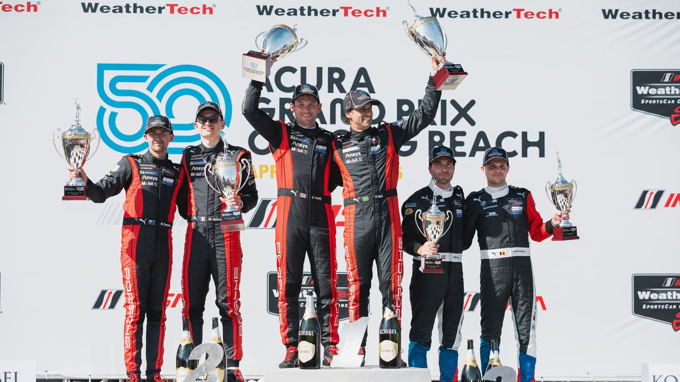 Race car drivers on the podium celebrate a thrilling one-two finish, clutching their trophies at the Acura Grand Prix of Long Beach while posing in front of a Porsche-branded backdrop.