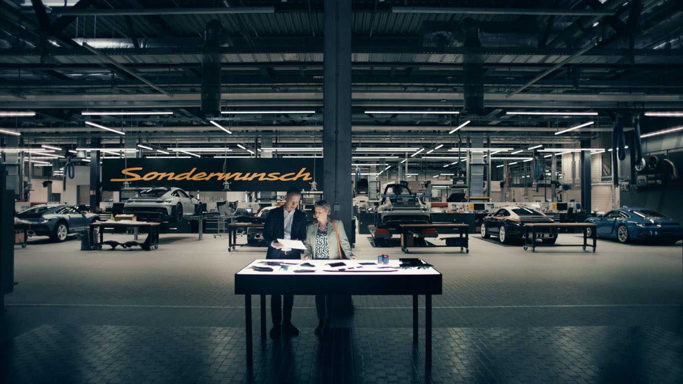 Two people stand at a table reviewing documents in a well-lit Porsche workshop, with several cars and a large "Sonderwunsch" sign in the background, capturing a behind-the-scenes moment from the video series.