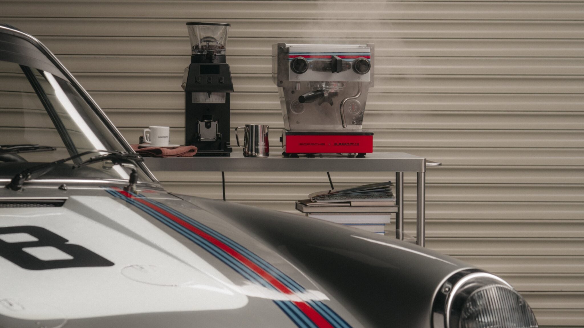 A sleek Porsche is parked in front of a table with a La Marzocco coffee machine emitting steam, a trusty coffee grinder, and books stacked cozily on one side.