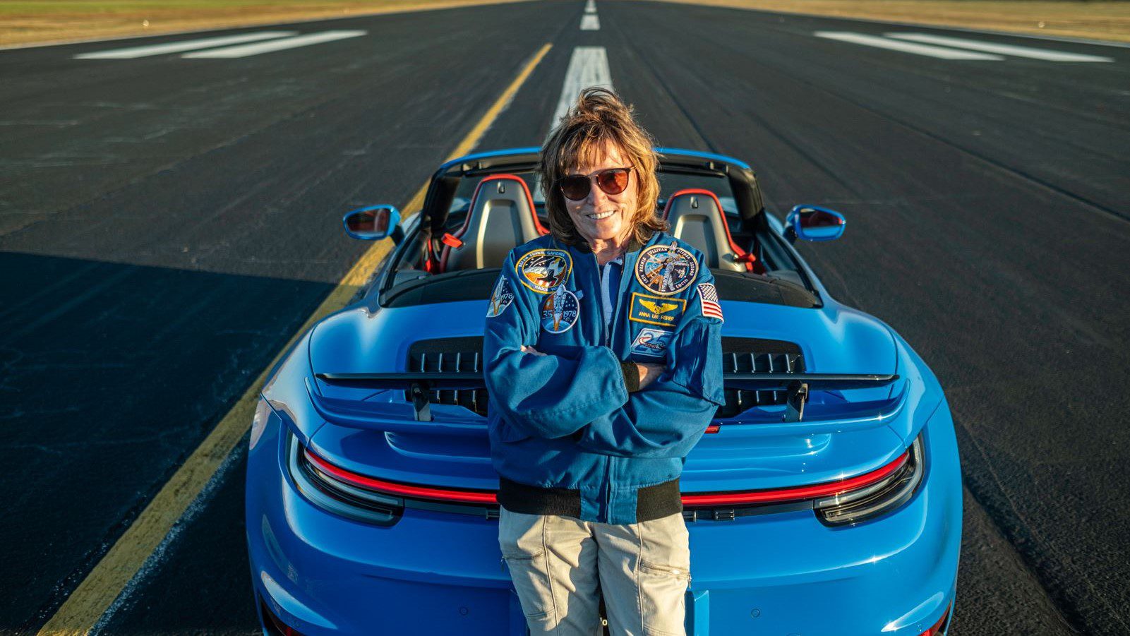 A person in a blue jacket and sunglasses stands with arms crossed, exuding confidence next to a blue convertible Porsche 911 Turbo parked on an airstrip, evoking the cool charisma of an astronaut prepping for launch.