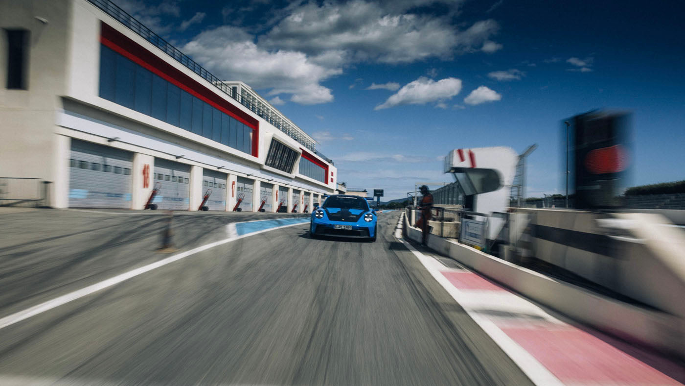 A blue Porsche zooms around the Circuit Paul Ricard, racing past the pit stop area under a blue sky with clouds. This thrilling track experience showcases the car's power and agility.