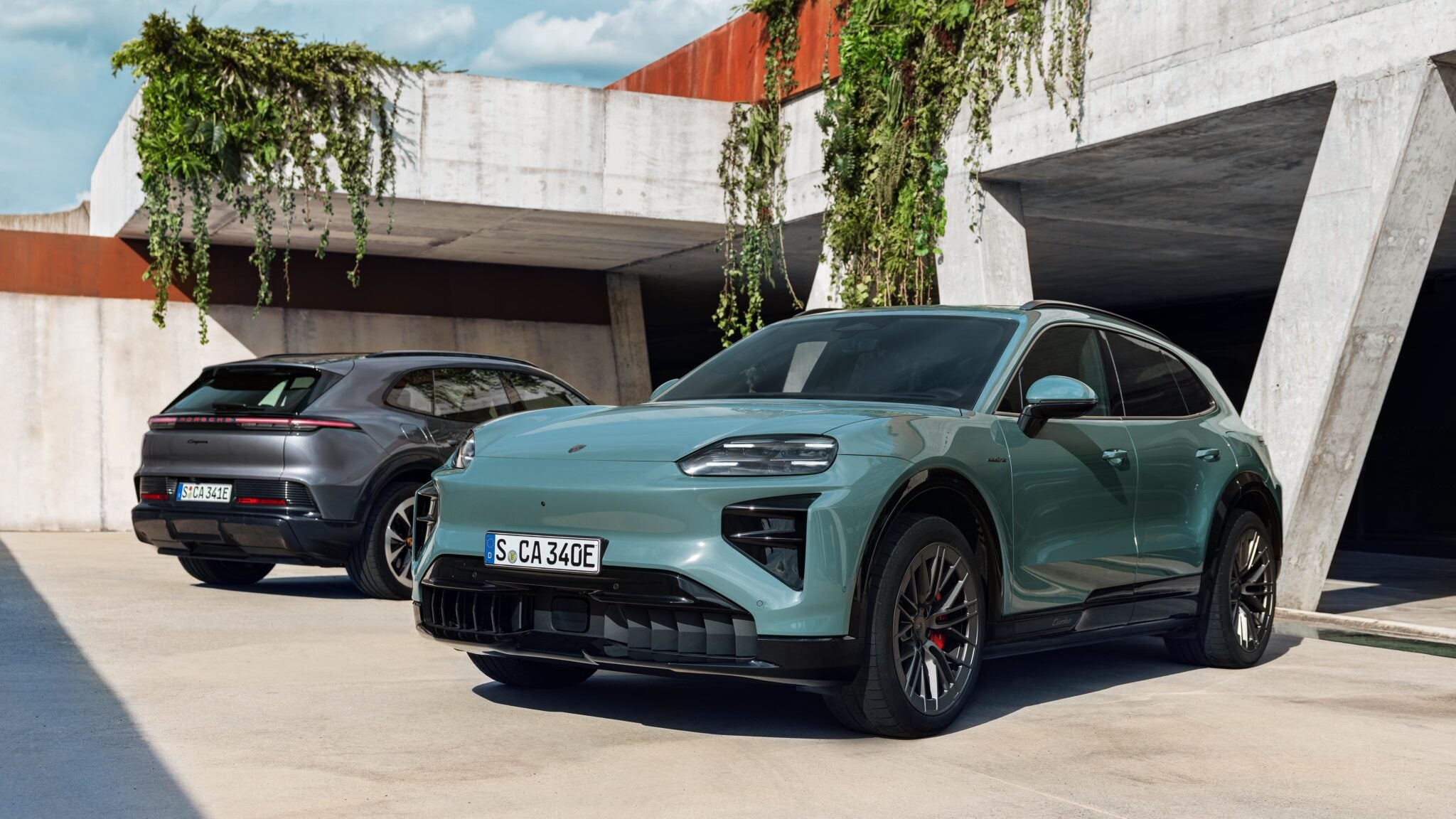 Two modern electric SUVs, including The Most Powerful Production Porsche Ever: 2026 Cayenne Electric, are parked outside a concrete building with plants hanging from above; both vehicles have European license plates.